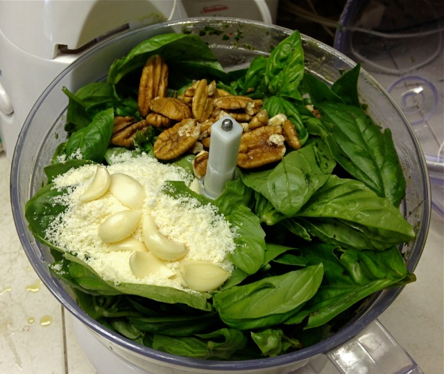 The food processor filled with basil, olive oil, parmesan cheese, walnuts and garlic